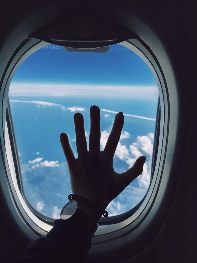 Home Silhouette of a hand touching an airplane window with a view of clouds and the sky.